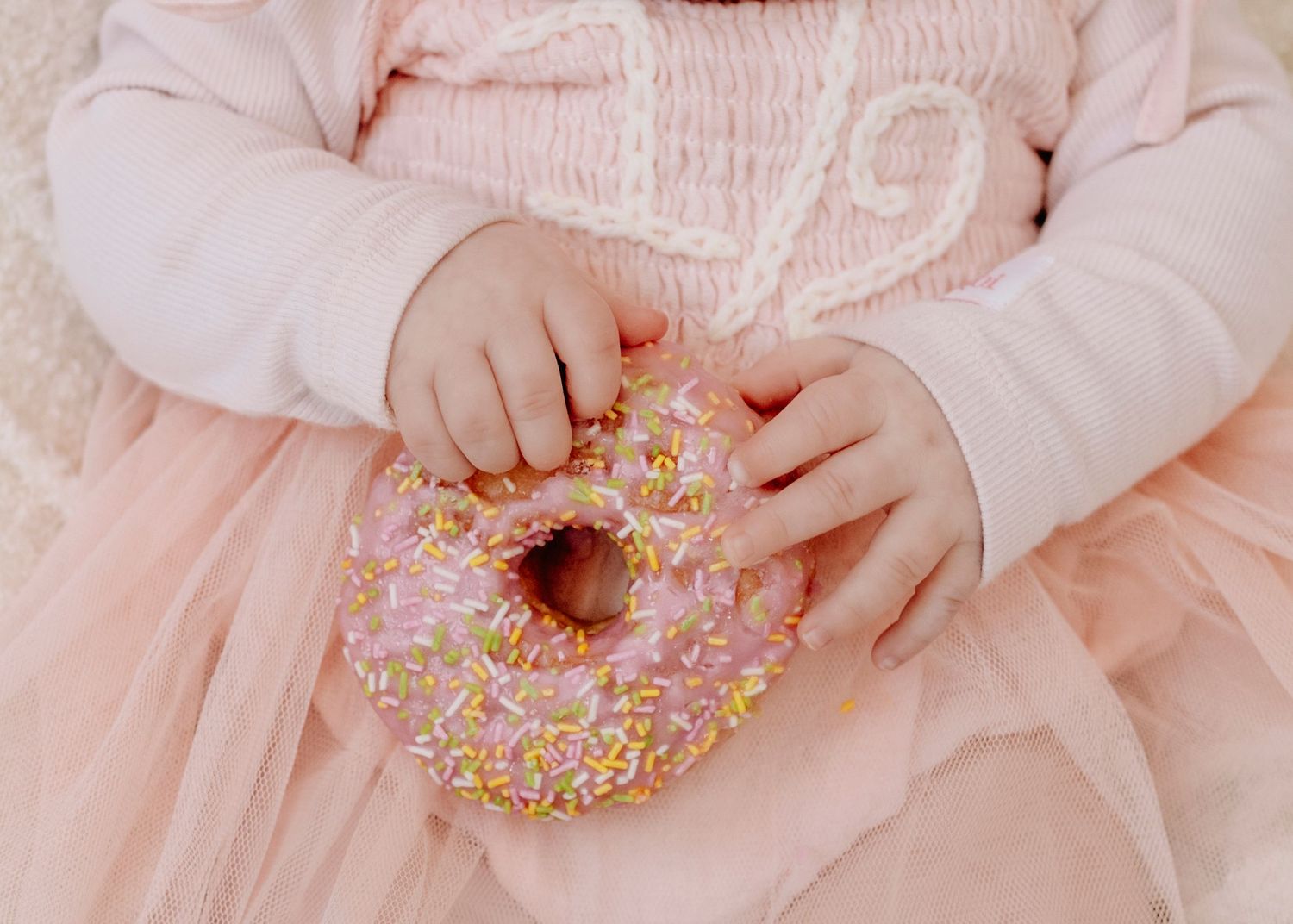 Small hands holding a pink frosted donut with sprinkles against a pink tulle dress.