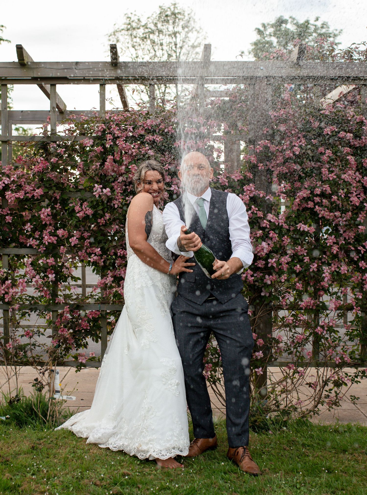 Newlyweds share a playful moment by a flowering garden wall.