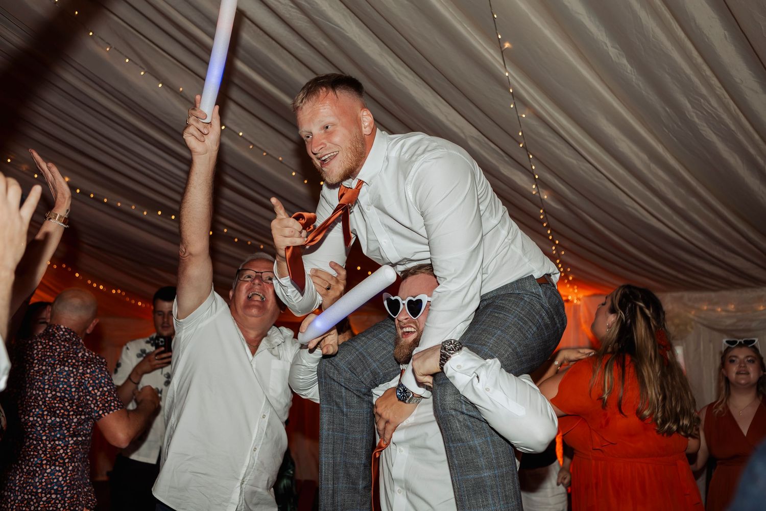 Wedding guests dancing and celebrating energetically on a dance floor with orange ambient lighting.
