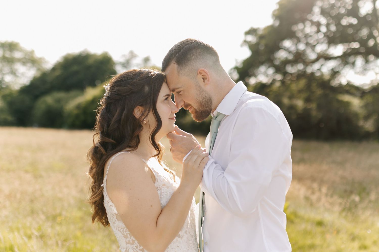 Intimate moment between couple in white attire standing in a sunlit meadow.