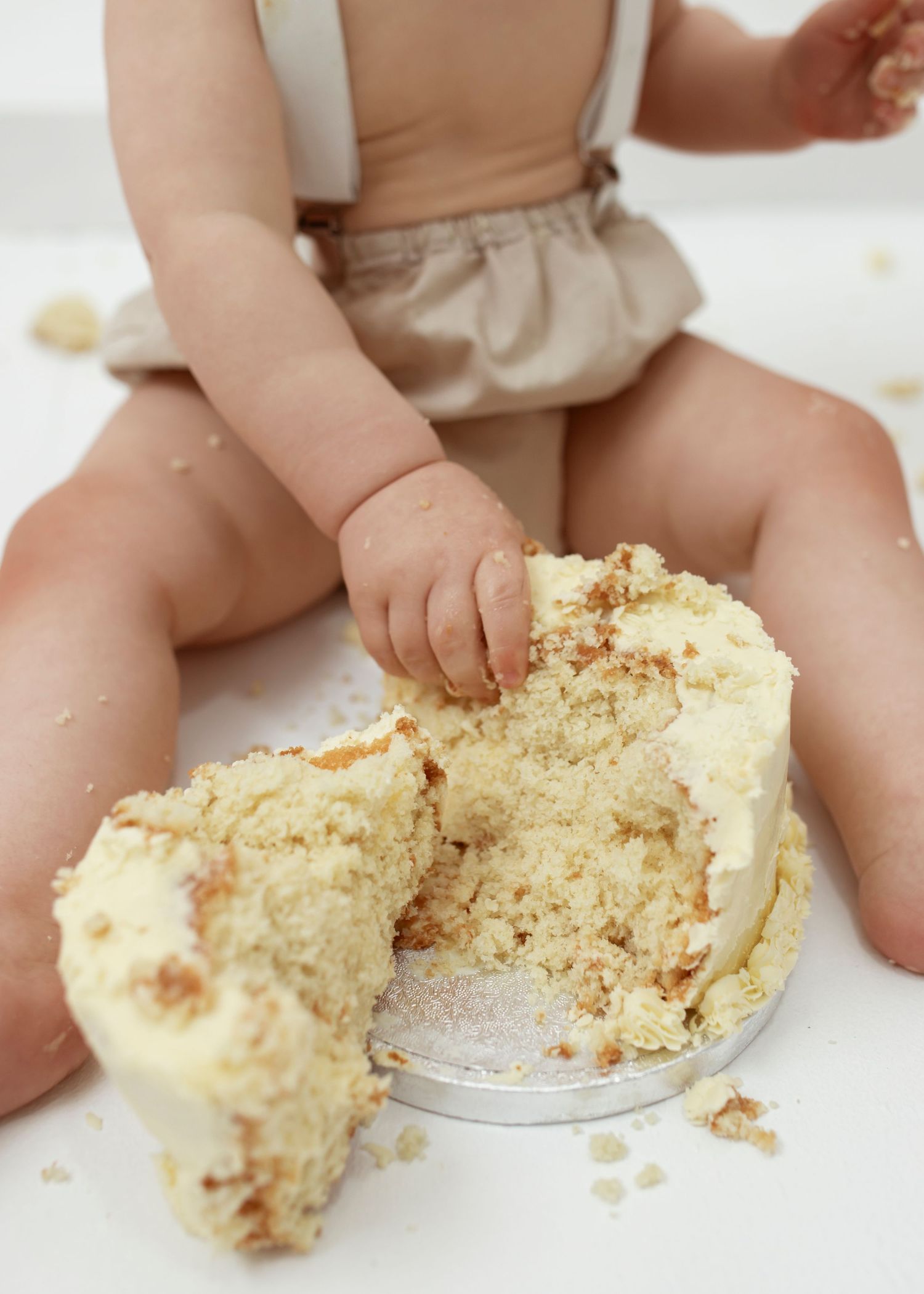 Baby in diaper plays with crumbled cake on white floor during cake smash photo session.