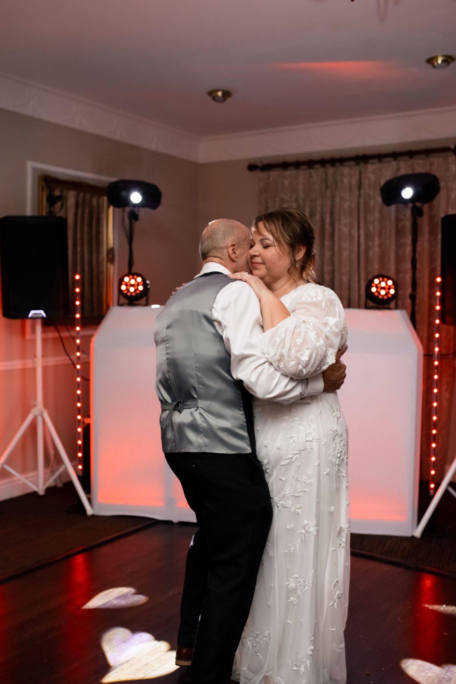 A couple shares a romantic dance in formal attire at a reception with red ambient lighting.
