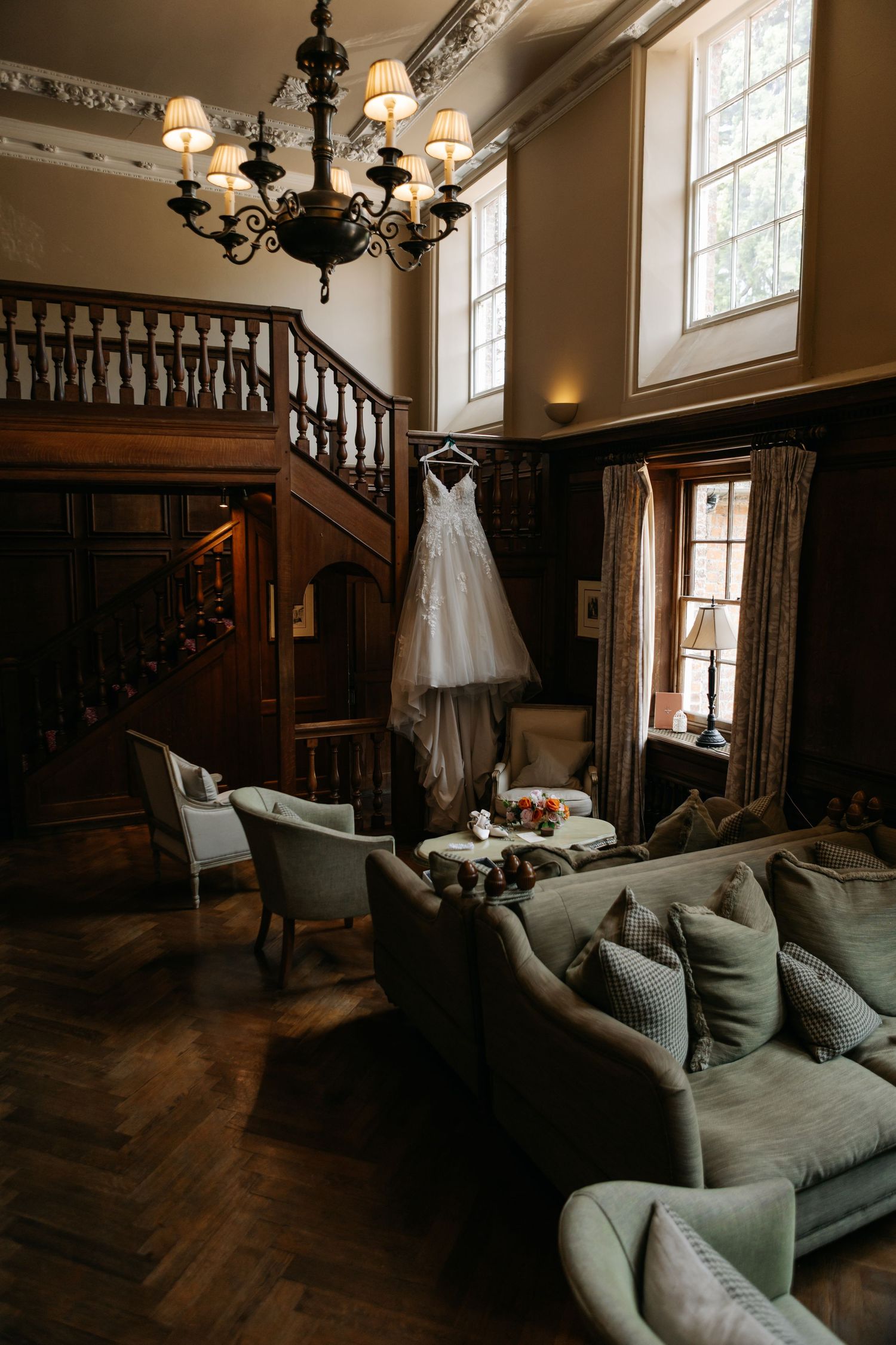 An elegant room with dark wood paneling, grey couch, and chandelier in warm lighting.