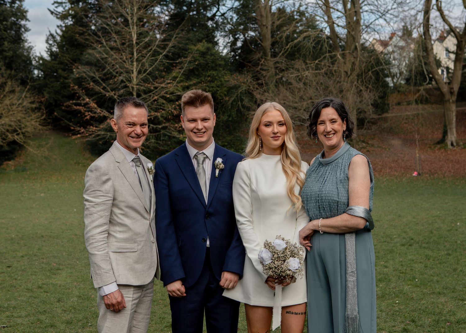 Small wedding party group poses together on grass with trees in background.