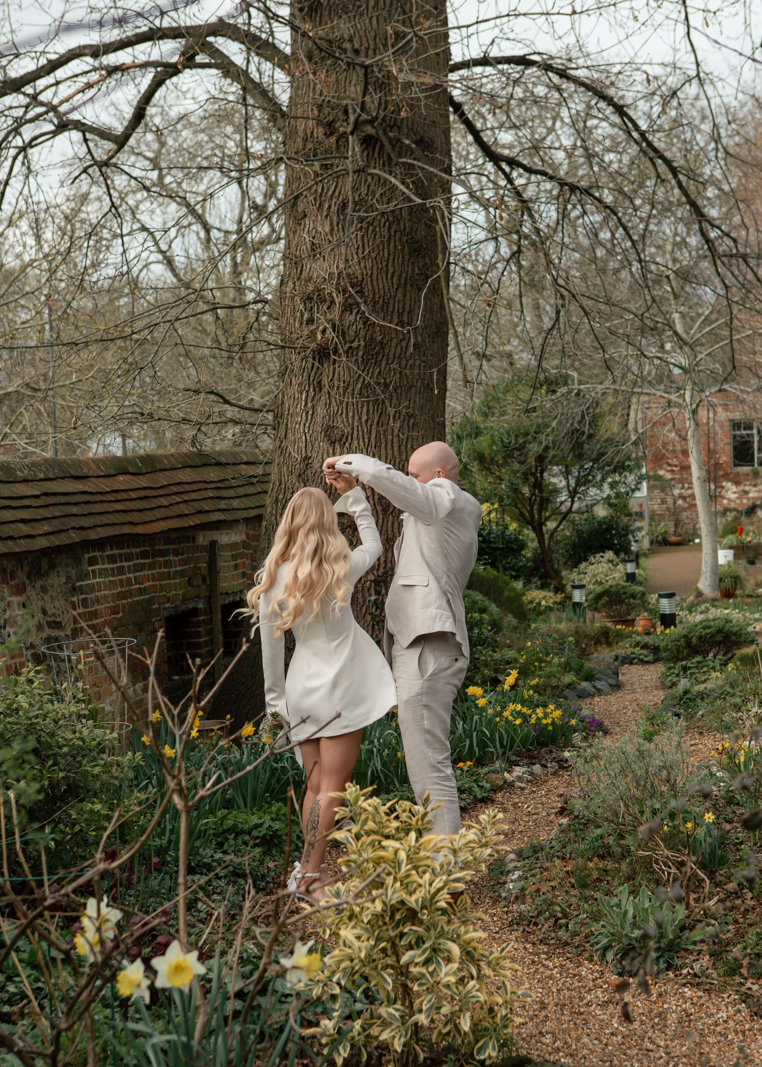 Dancing couple in coordinated white outfits surrounded by spring flowers and garden landscaping.