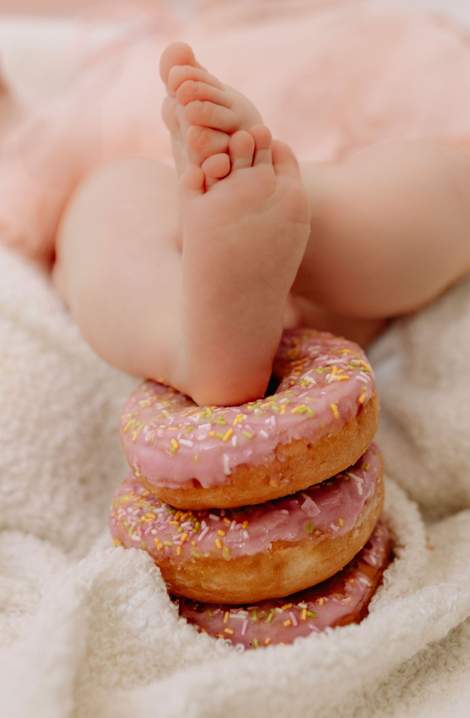 Tiny baby foot resting on stack of pink frosted donuts in artistic photography setup.