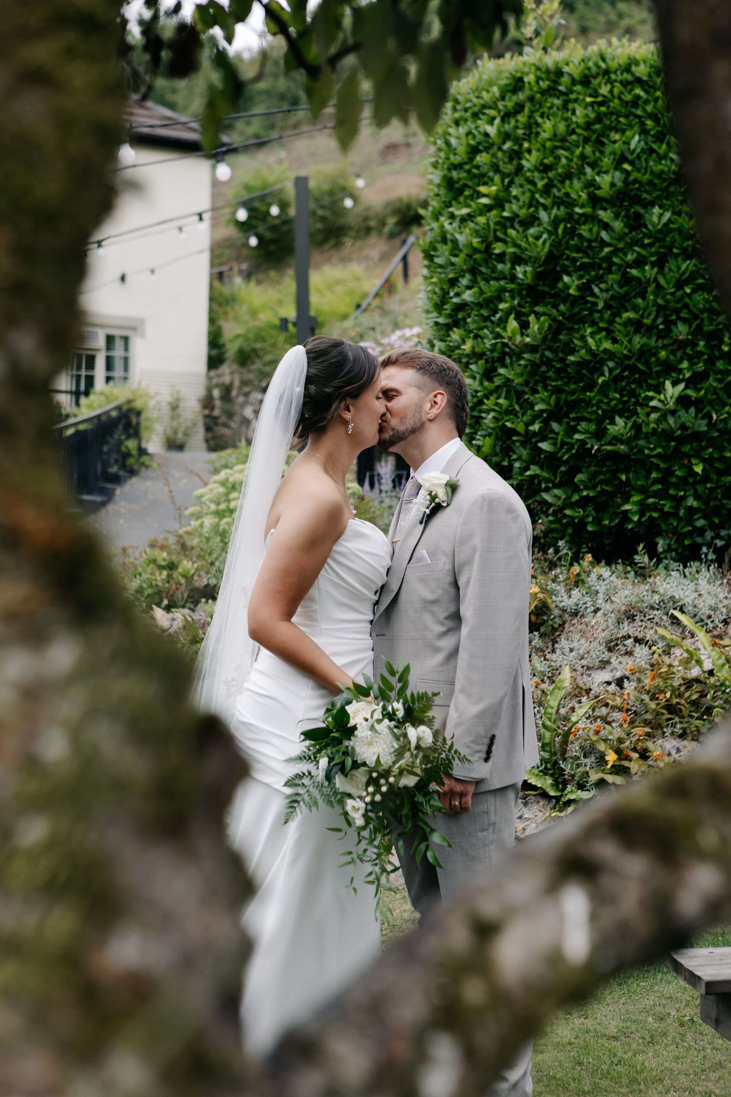 Newlyweds share tender moment framed by garden foliage.
