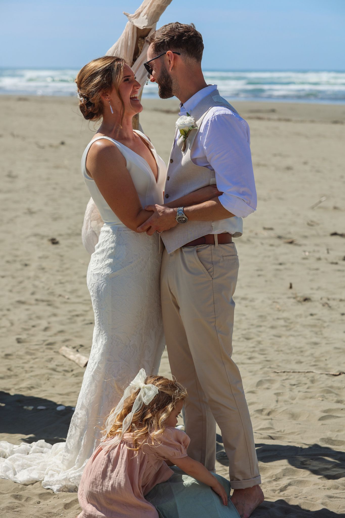 Newlyweds look at each other and smile after their first kiss as their daughter sits by their feet playing with shells in the sand.
