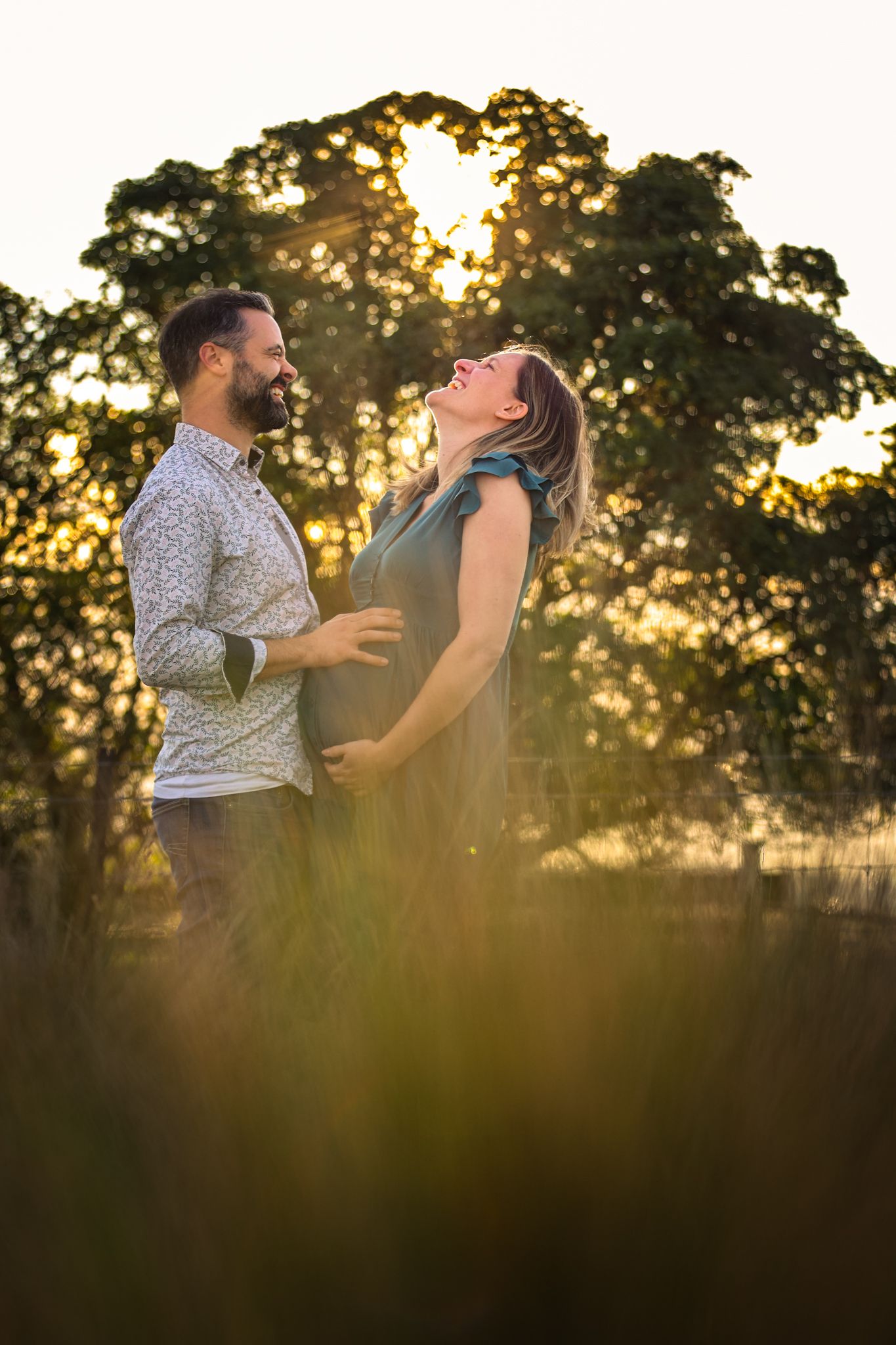 Expecting couple laughing in front of a tree with golden light peaking through during their maternity photoshoot