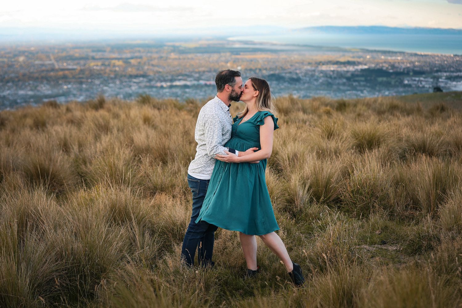Expecting couple having a maternity photo shoot on the Christchurch Port Hills, standing in a field of long grass with the city scape behind