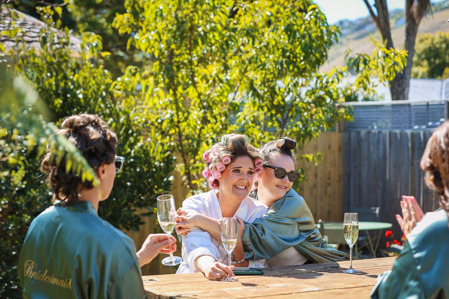 Bride sitting with her bridesmaids, sipping champagne in silk dressing gowns and curlers on the morning of the wedding