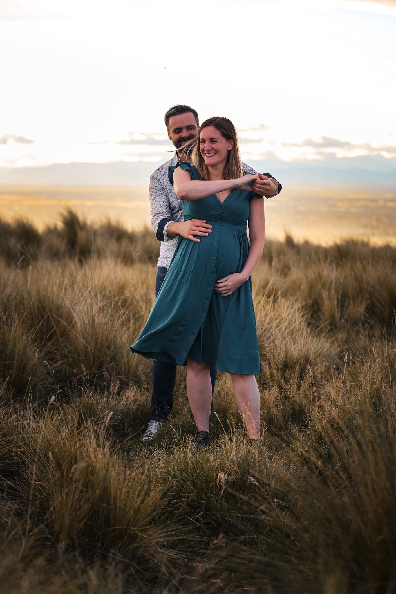 Expecting couple dancing at their maternity shoot on the Christchurch Port Hills in the long grass with city scape behind them