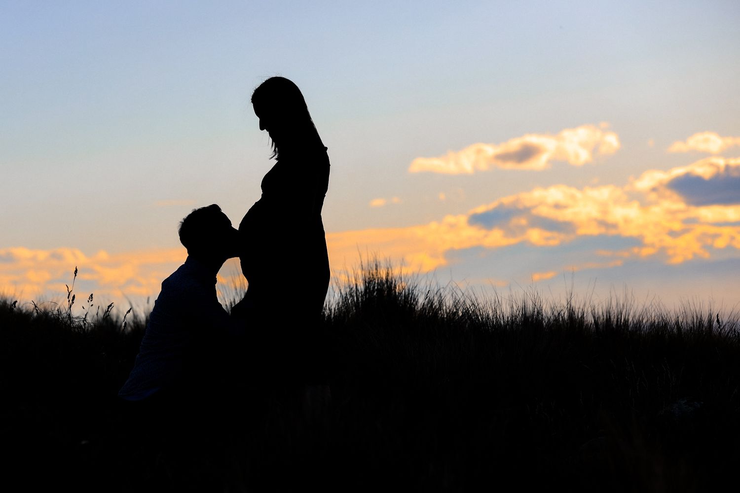 Silhouette of an expecting couple, man kissing pregnant stomach against a golden sunset.
