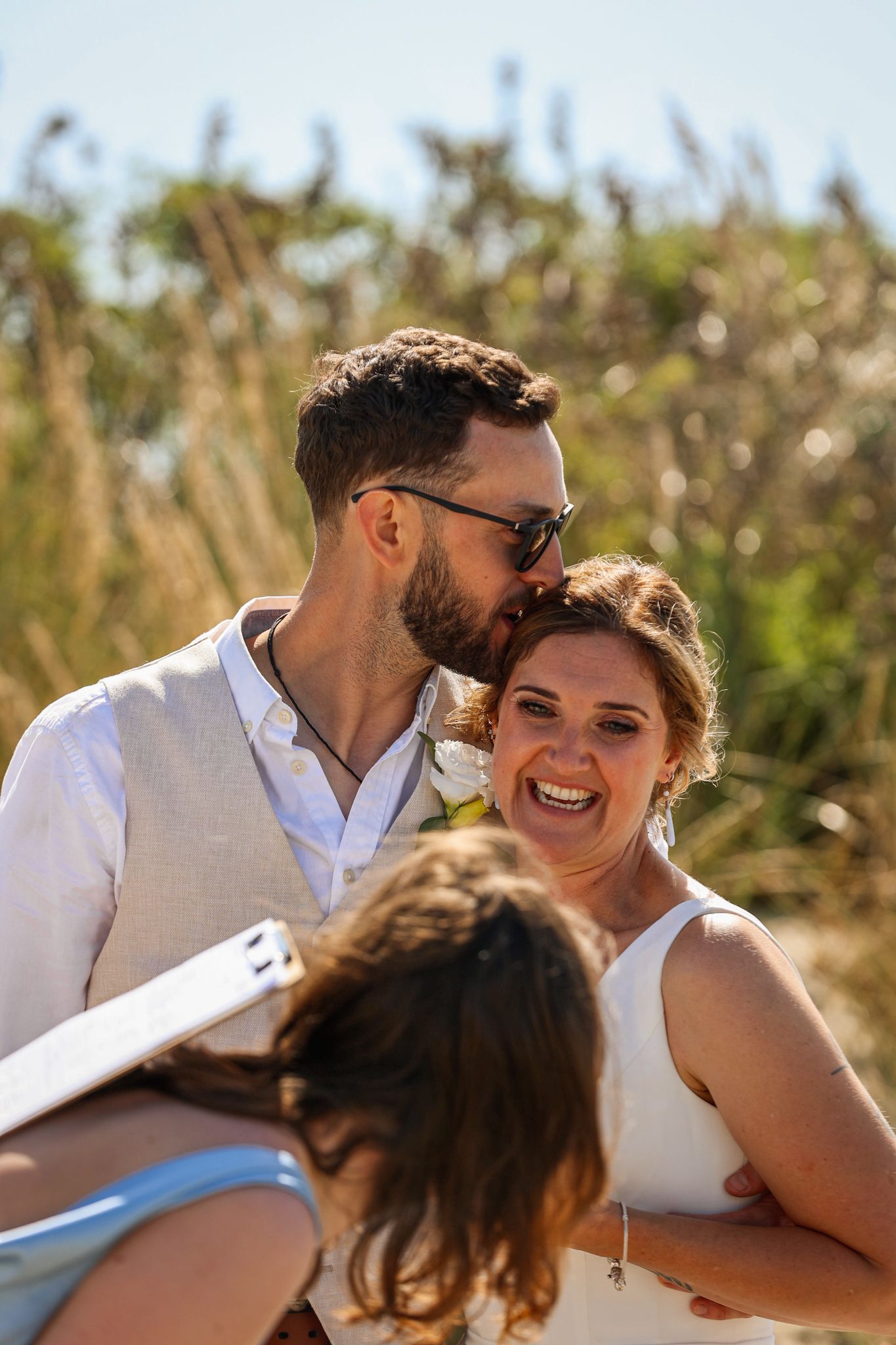 Groom kisses bride on forehead while witness signs marriage documents