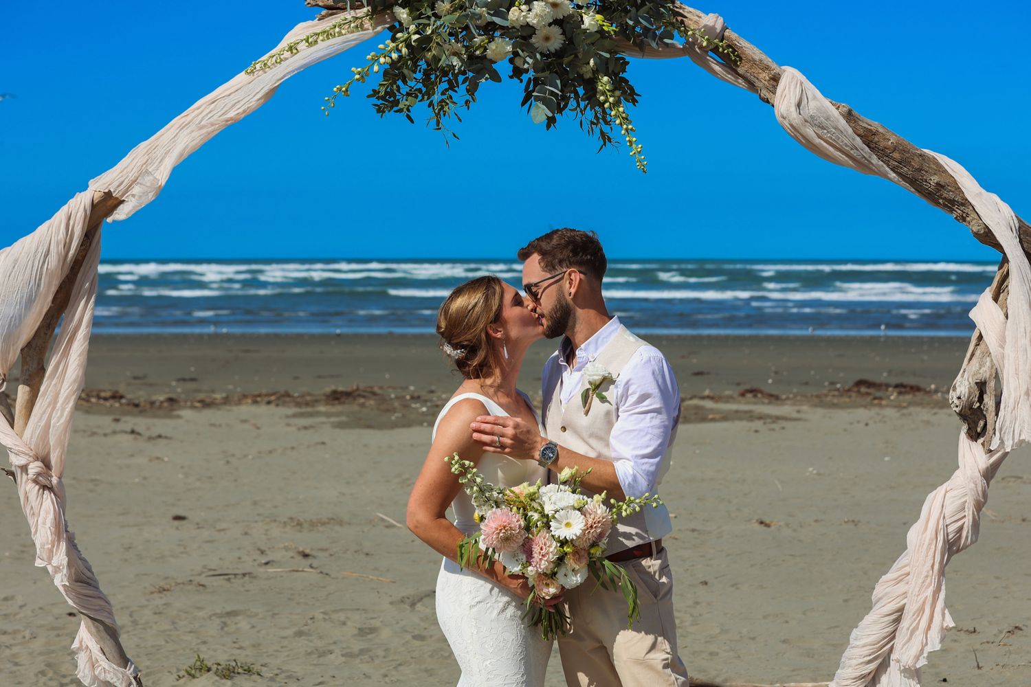 Newlyweds caught in their first kiss beneath a beachside wedding arch