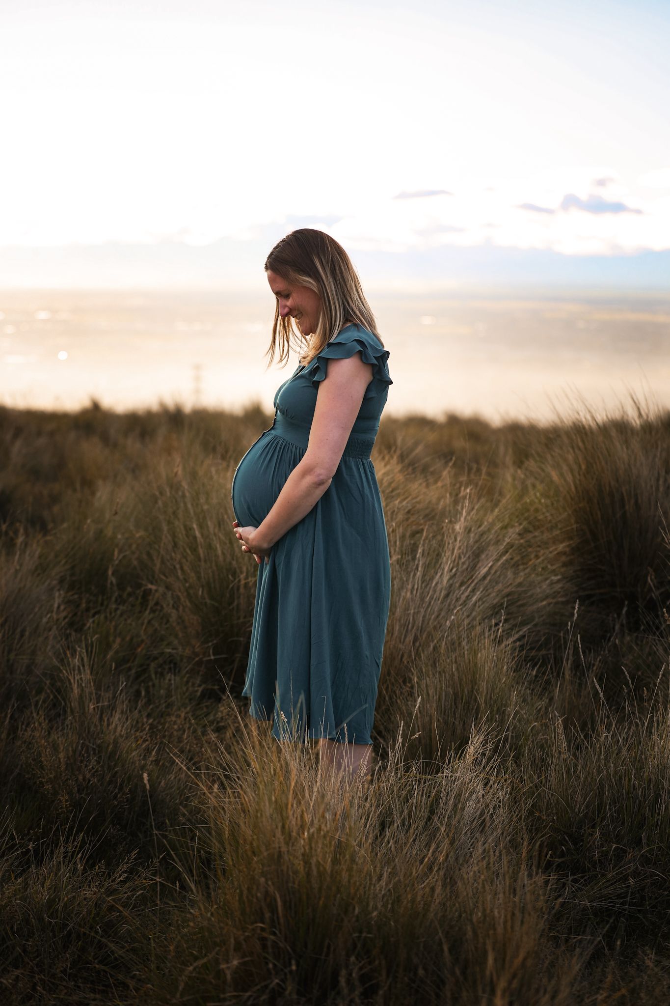 Expecting mother holding her belly standing in the long grass during her maternity photoshoot at sunset in christchurch port hills