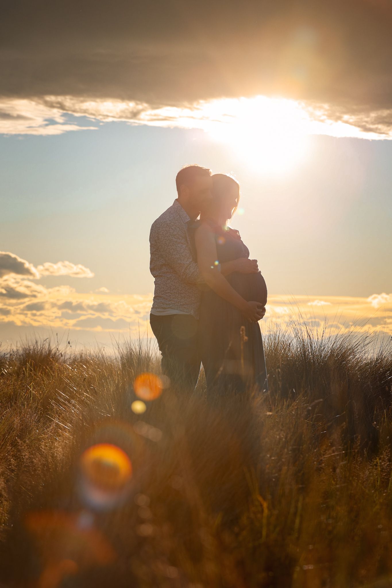 Lens flare peeking through the clouds in front of an expecting couple during their maternity photoshoot