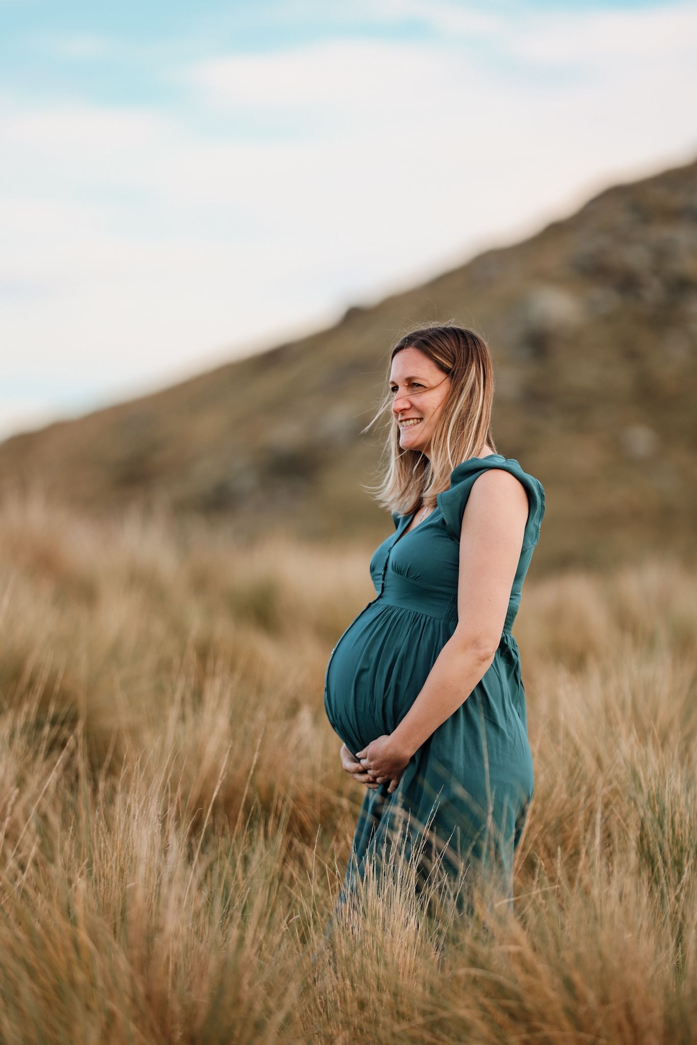 Expecting mother holding her belly standing in the long grass during her maternity photoshoot at sunset in christchurch port hills