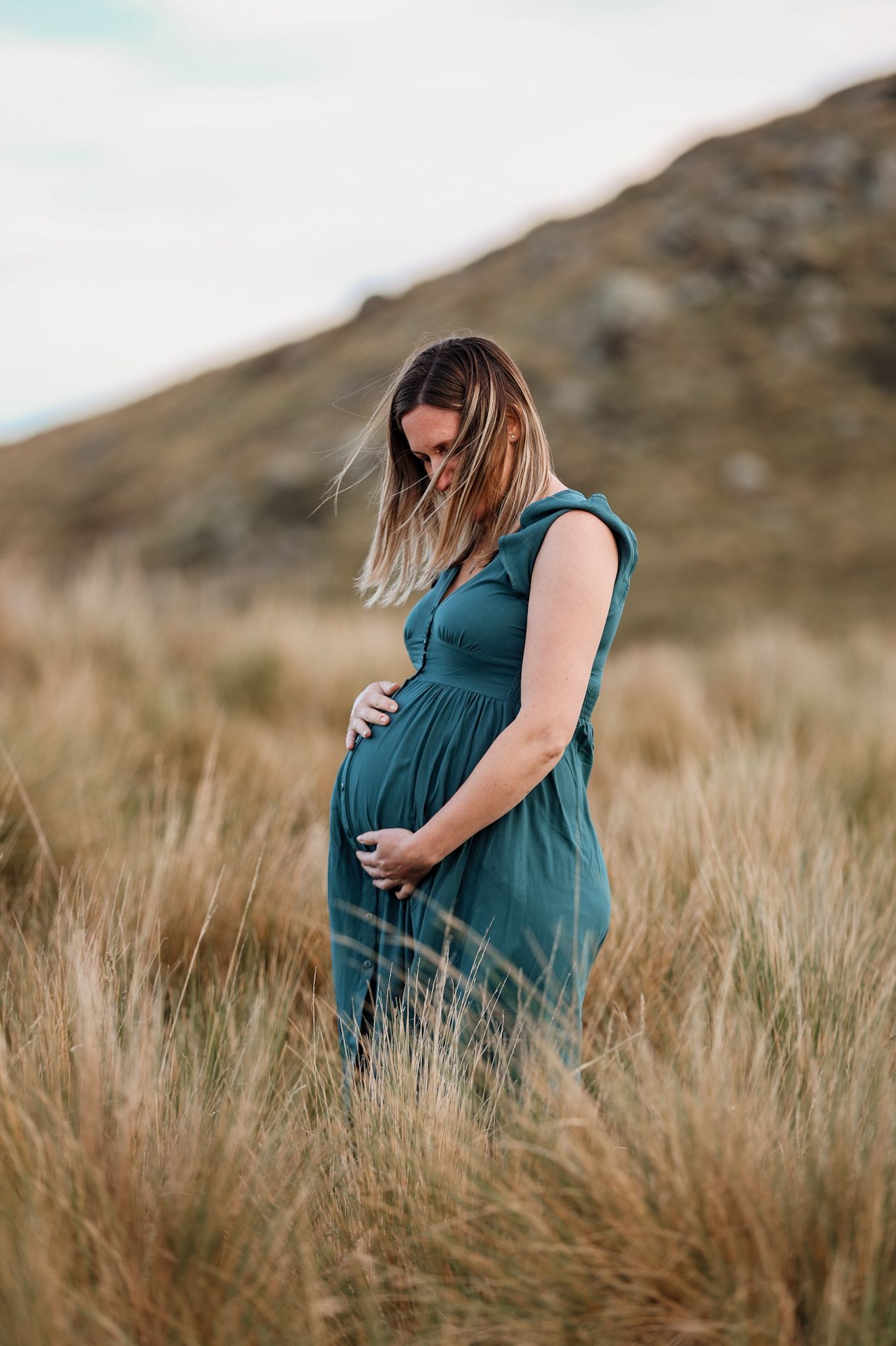Expecting mother holding her belly standing in the long grass during her maternity photoshoot at sunset in christchurch port hills