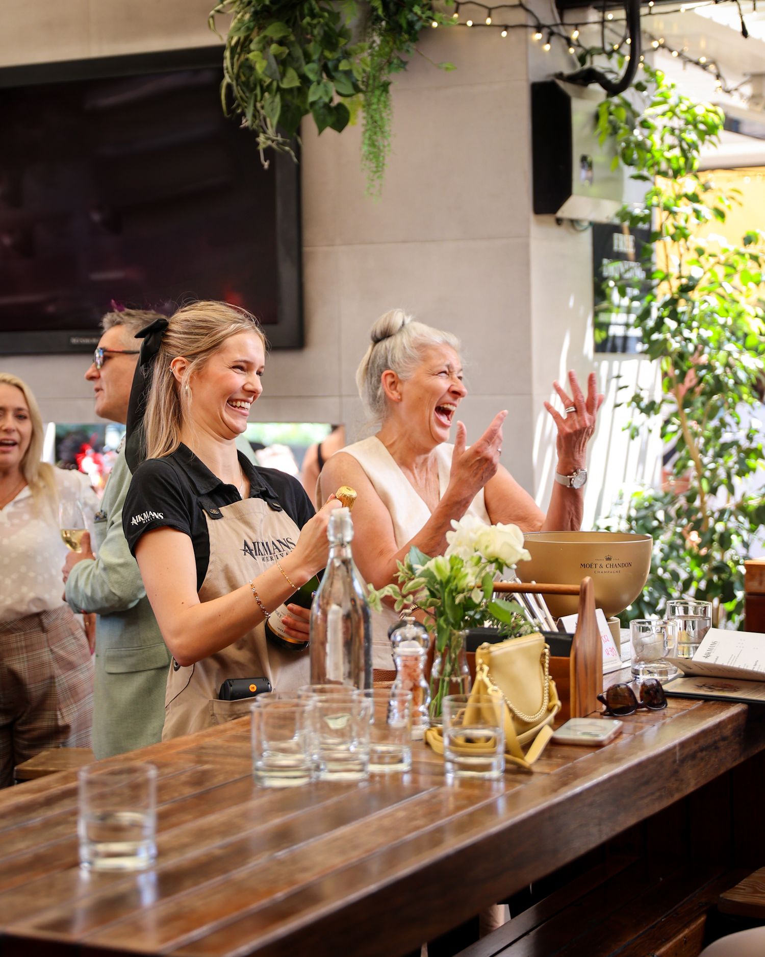 Waitress and customer laughing as she opens a bottle of champagne.