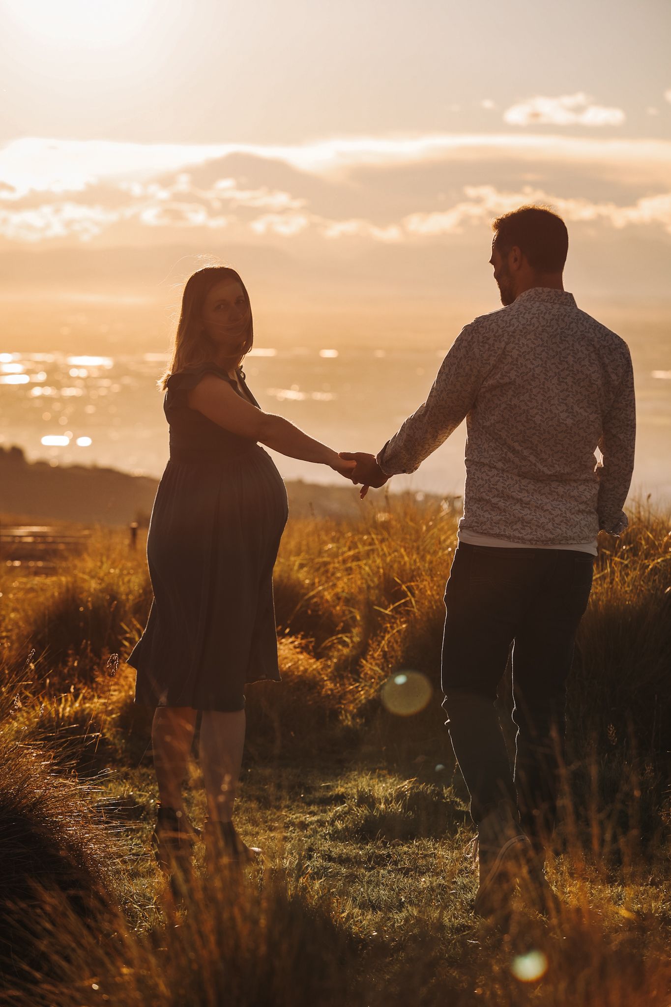 Expecting lady looks back towards the camera during her maternity photoshoot. Man holds her hand and looks off into the sunset.