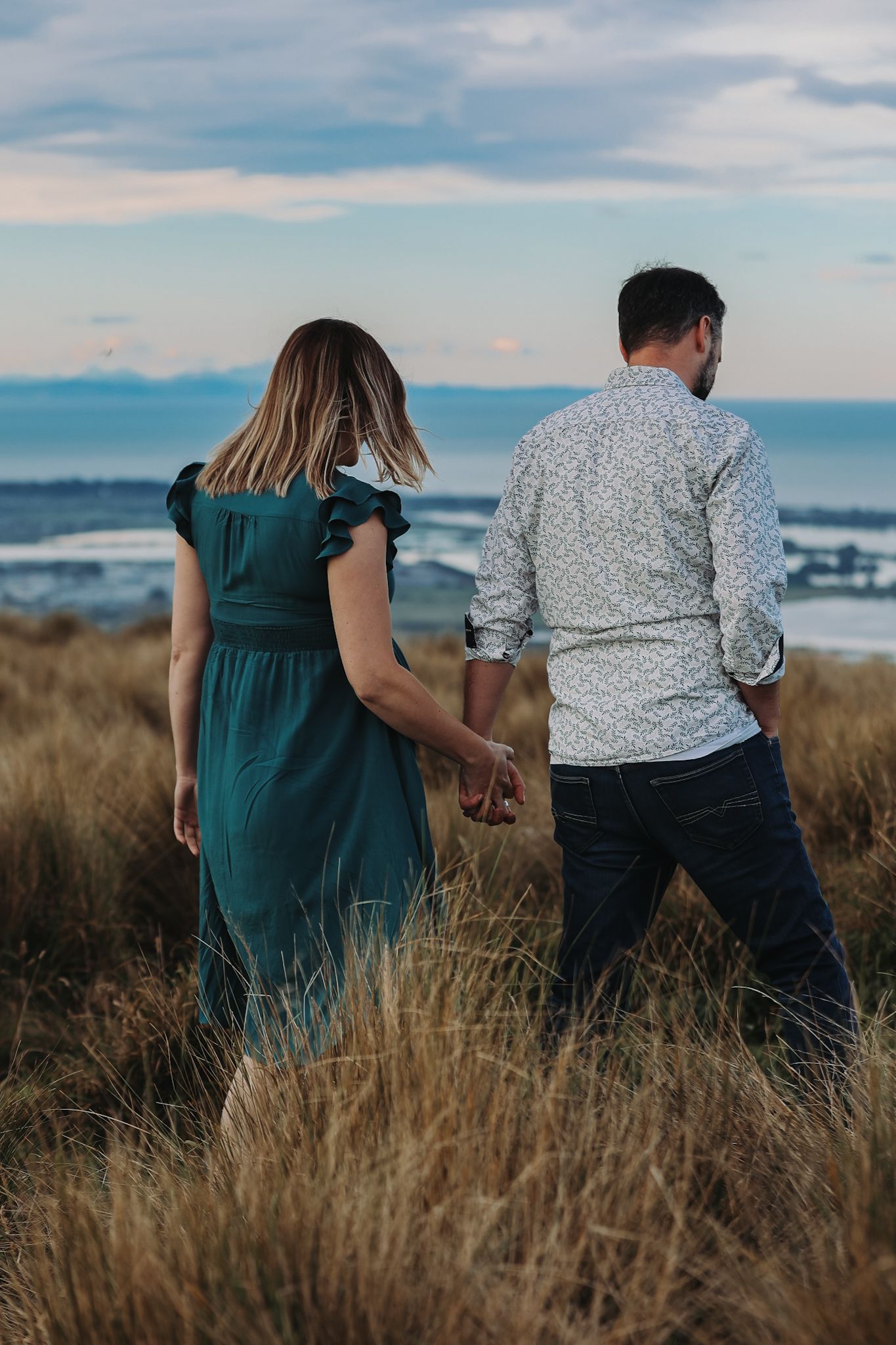 Expecting couple walk away from view during a maternity photoshoot against a view of the ocean from the christchurch port hills.