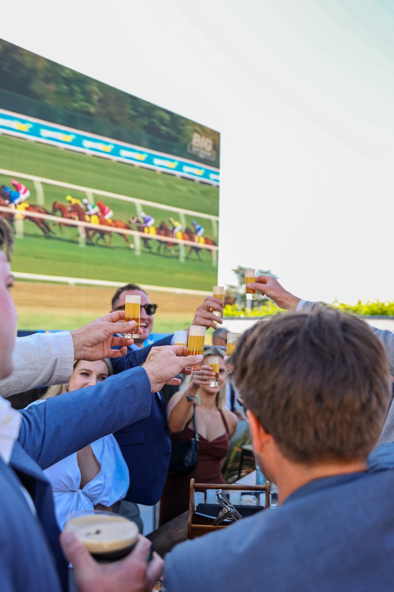 Group of friends cheersing a round of shots at Aikmans Merivale Cup Day event in front of a large TV screen showing the races.