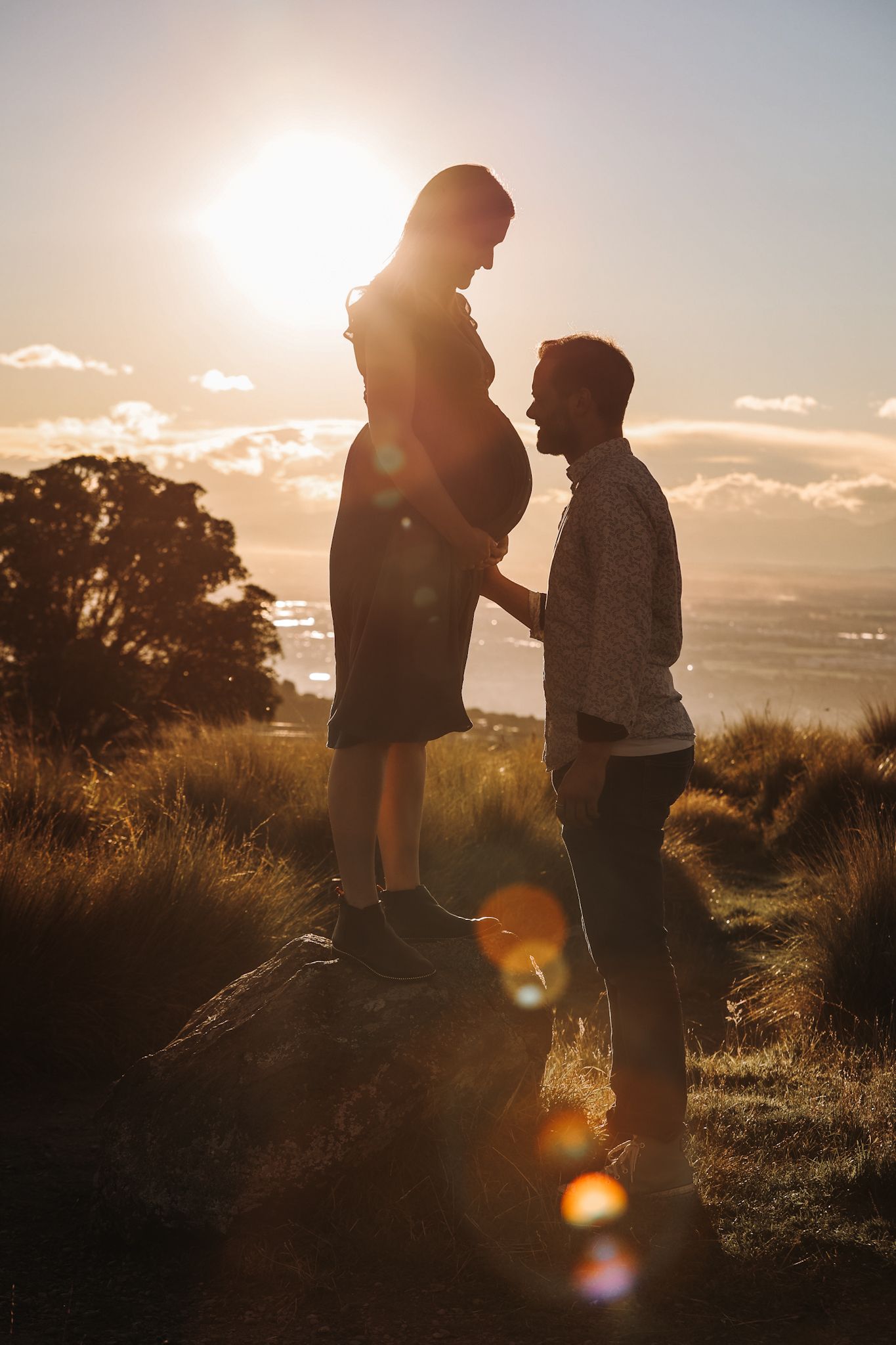 Pregnant woman standing on a rock at sunset, holding hands with her partner for maternity photos