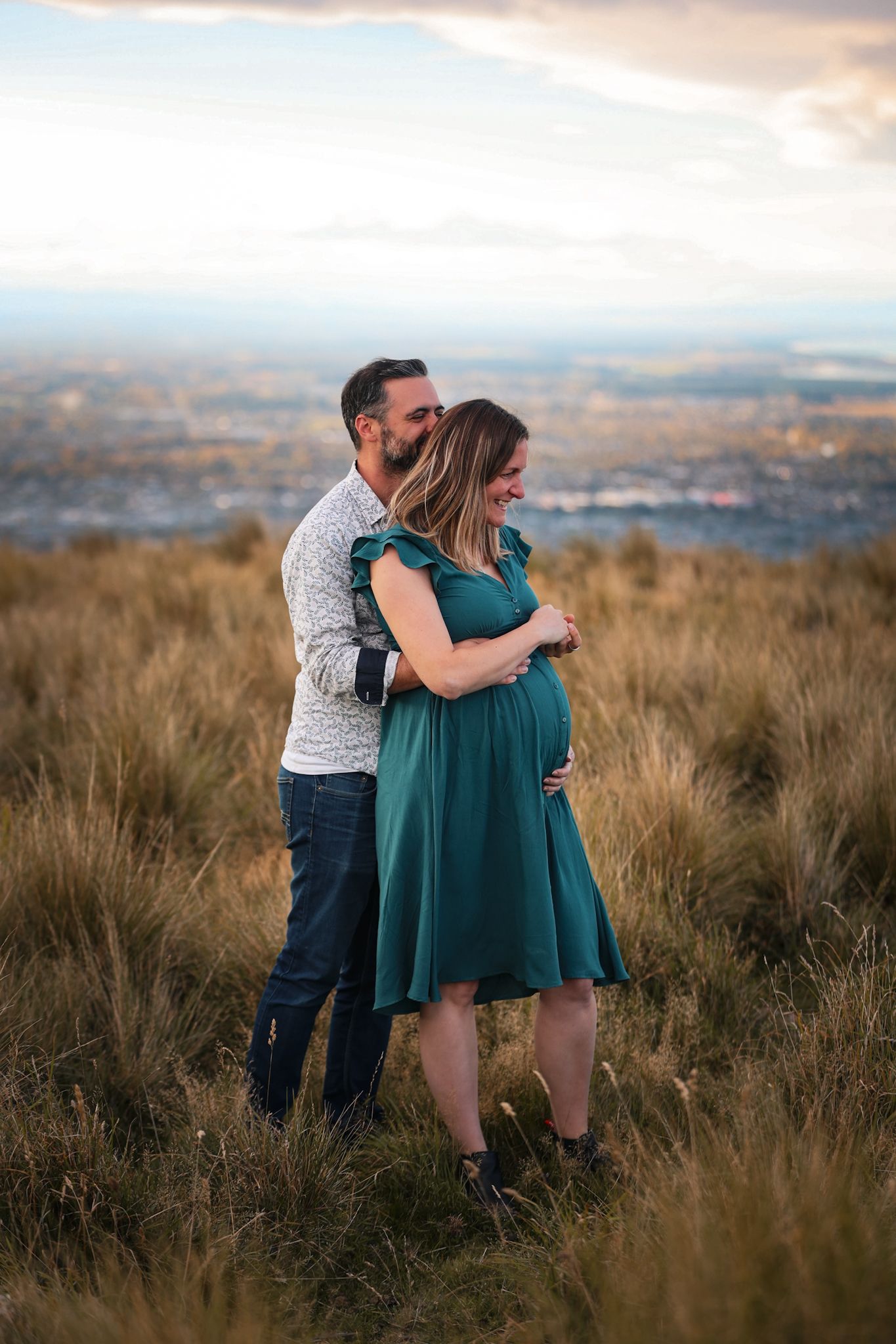 Expecting couple during a maternity photoshoot looking out onto christchurch city from the Port Hills whilst twirling in dance.