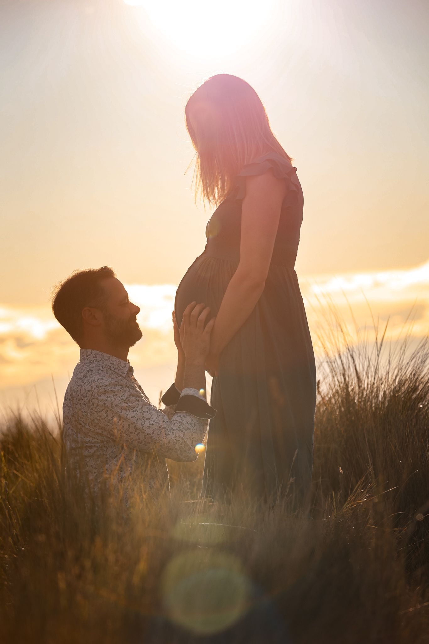 Lens flare peeking through the clouds in front of an expecting couple during their maternity photoshoot for golden hour