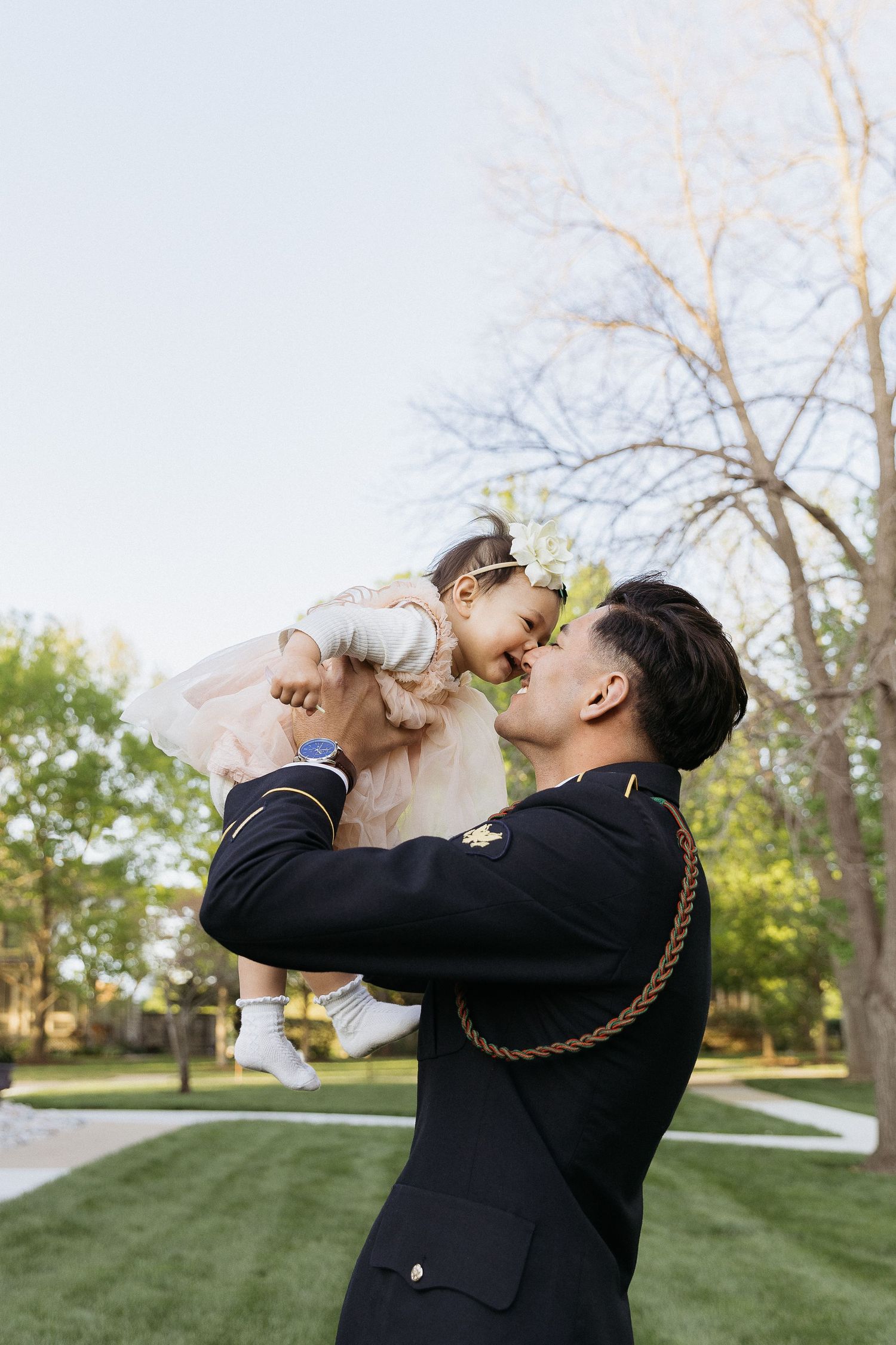 A serviceman in dress uniform lifts a small child in a light dress outdoors.