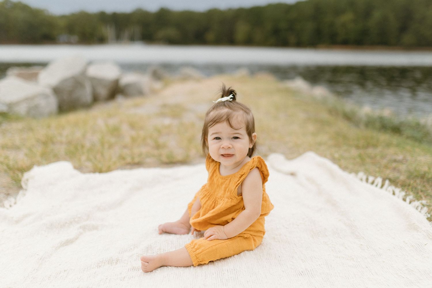 Small child in orange romper sits happily on sandy ground near water.