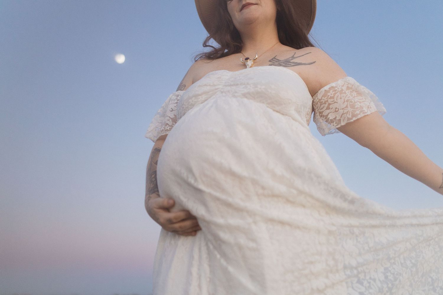 Close-up maternity portrait in white lace dress against sunset sky with visible moon.