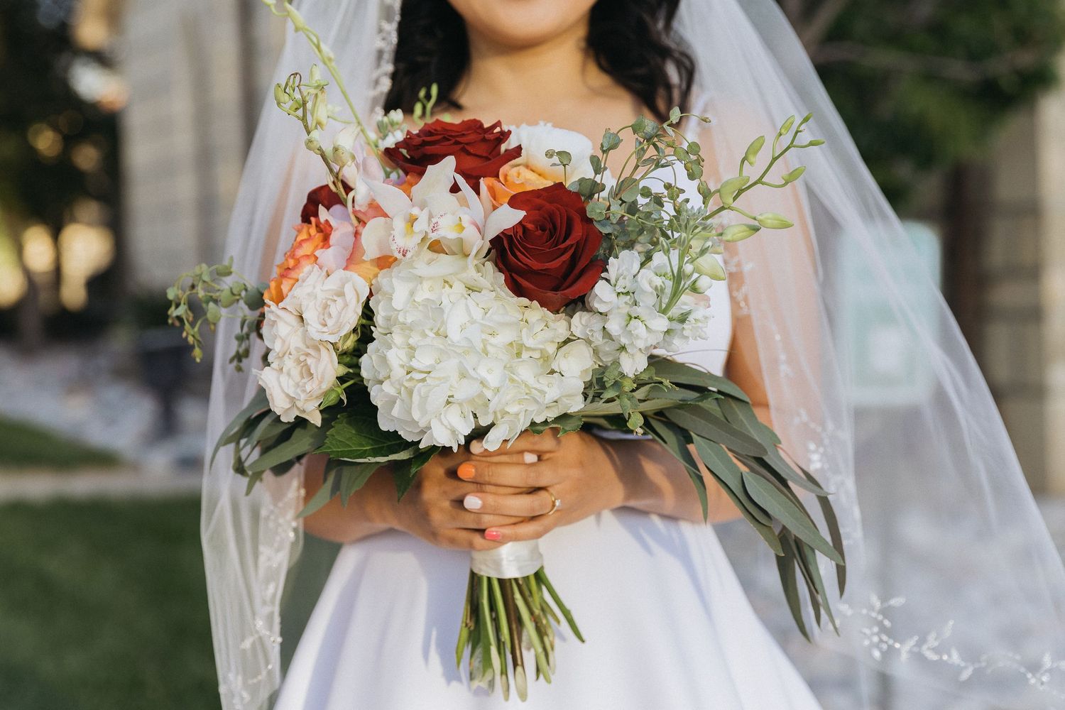 Elegant bridal bouquet with red roses, white hydrangeas and greenery held by bride in white dress.