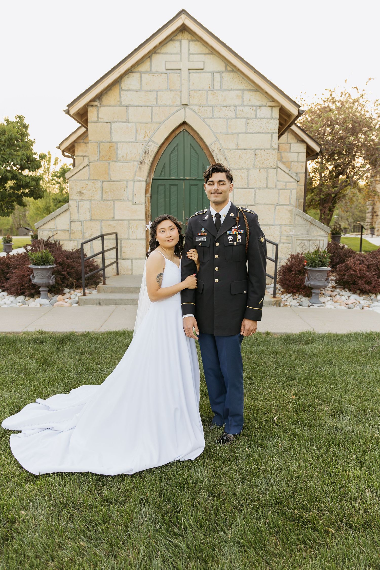 A wedding couple poses in front of a small stone chapel with a cross.