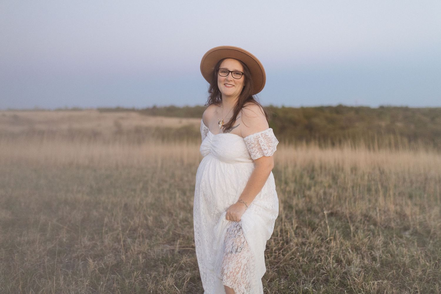 Casual maternity portrait in white off-shoulder dress and brown hat in field at dusk.