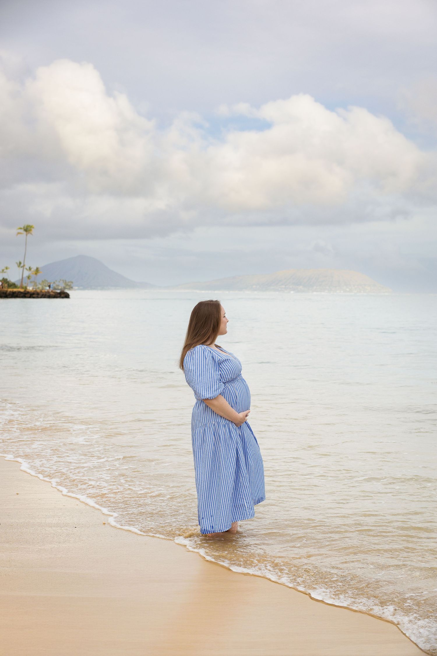 A person in a flowing light blue dress stands at the ocean's edge as gentle waves lap at the sandy shoreline.
