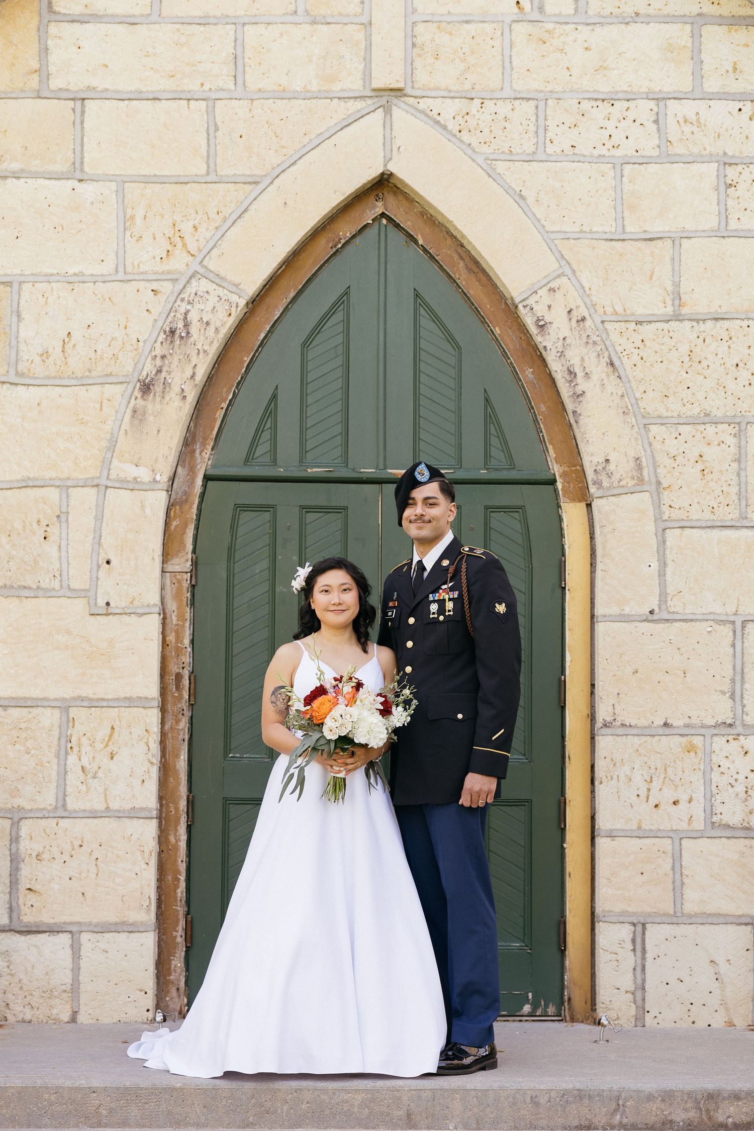 Wedding couple poses in front of arched green church door in formal military and bridal attire.