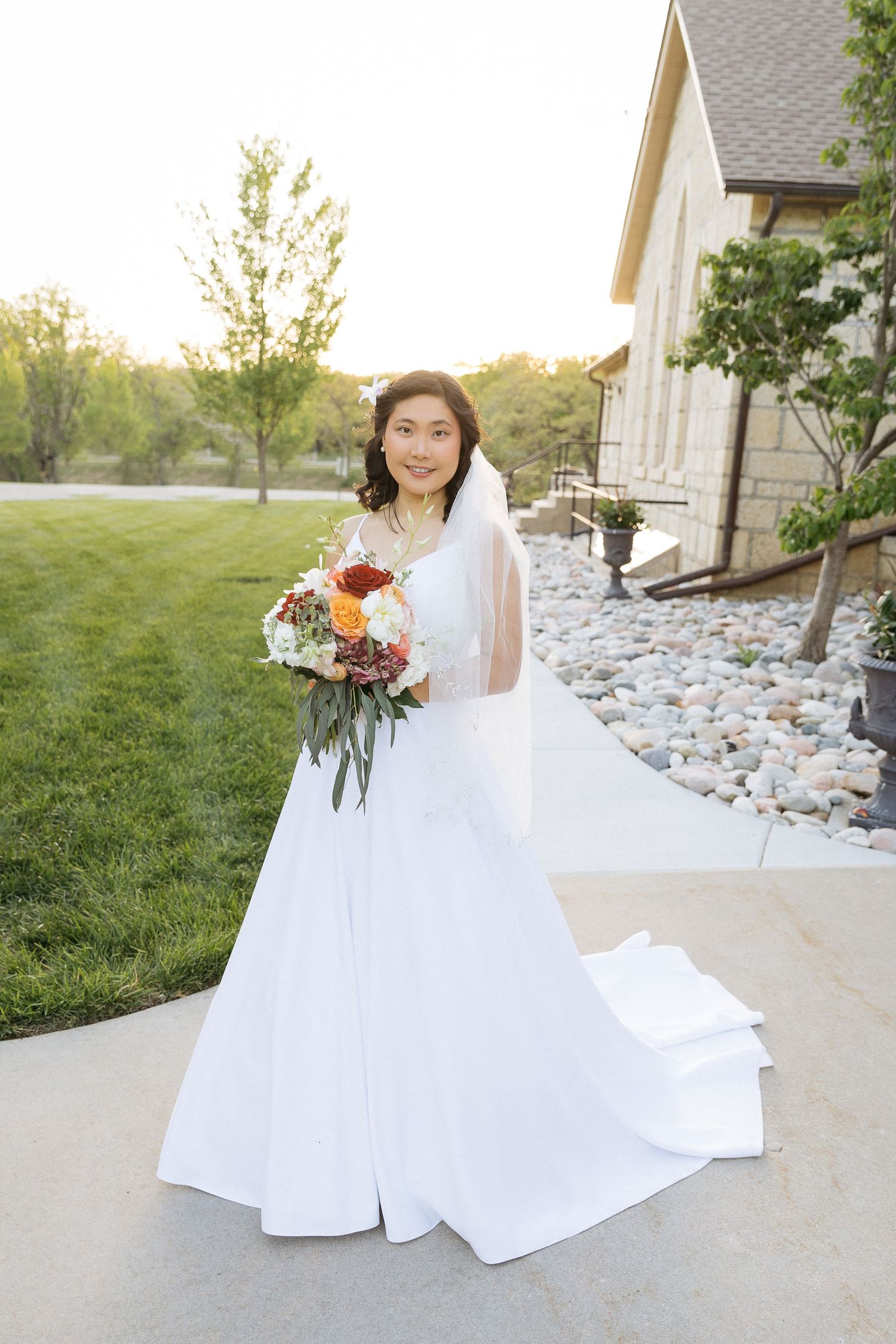 A bride in a flowing white wedding gown holds a vibrant fall bouquet while standing on a stone pathway at sunset.