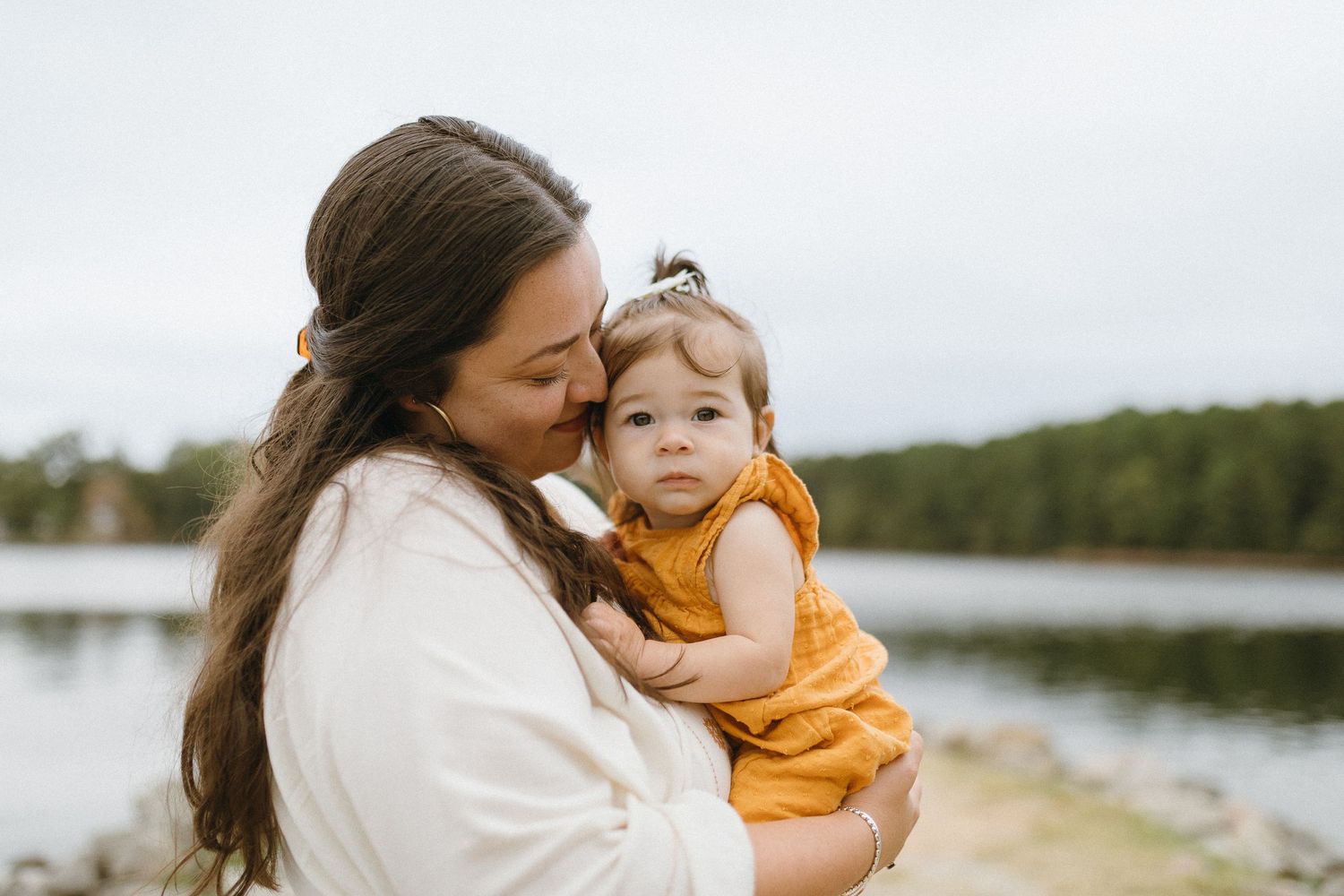 Mother in white dress holds child in orange outfit against lakeside backdrop.