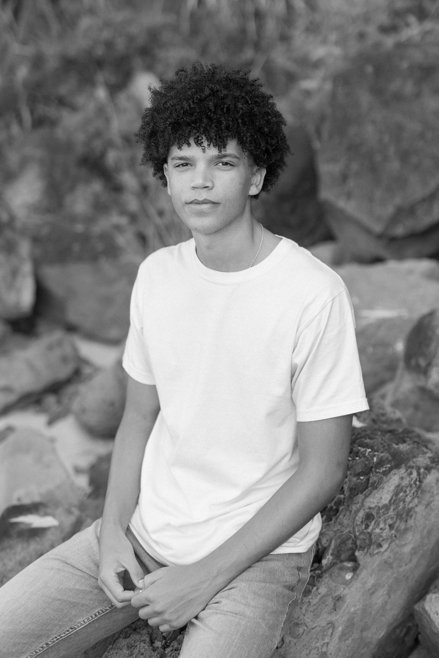 Black and white portrait of a young person in a white t-shirt sitting on rocks outdoors.  waimea bay senior session north shore oahu hawaii