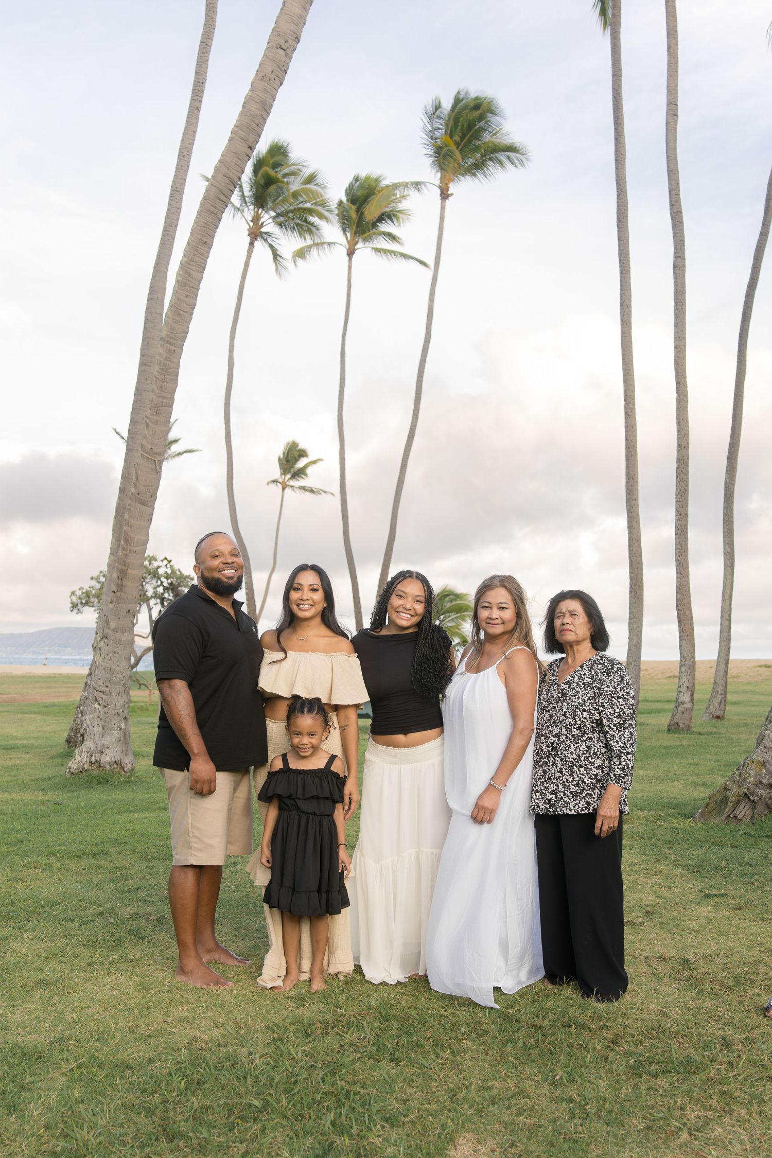 A group stands together on a grassy hill with tall palm trees swaying in the wind against a cloudy sky.