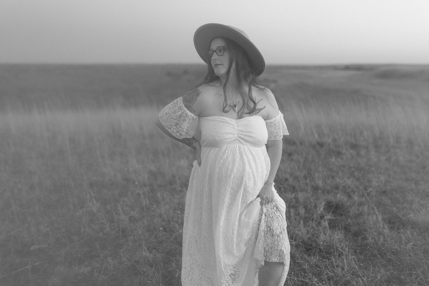 Black and white maternity photo featuring white dress and hat in rustic field setting.