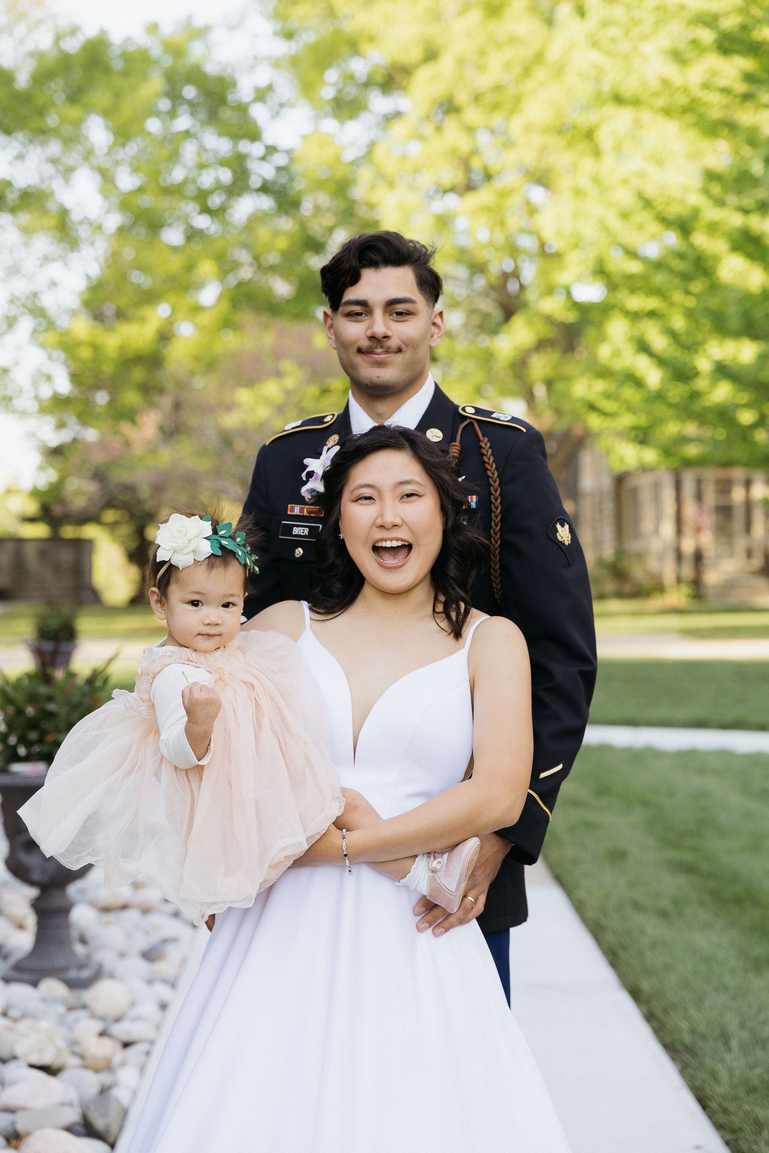 A joyful wedding portrait outdoors with couple and small child in pink dress.