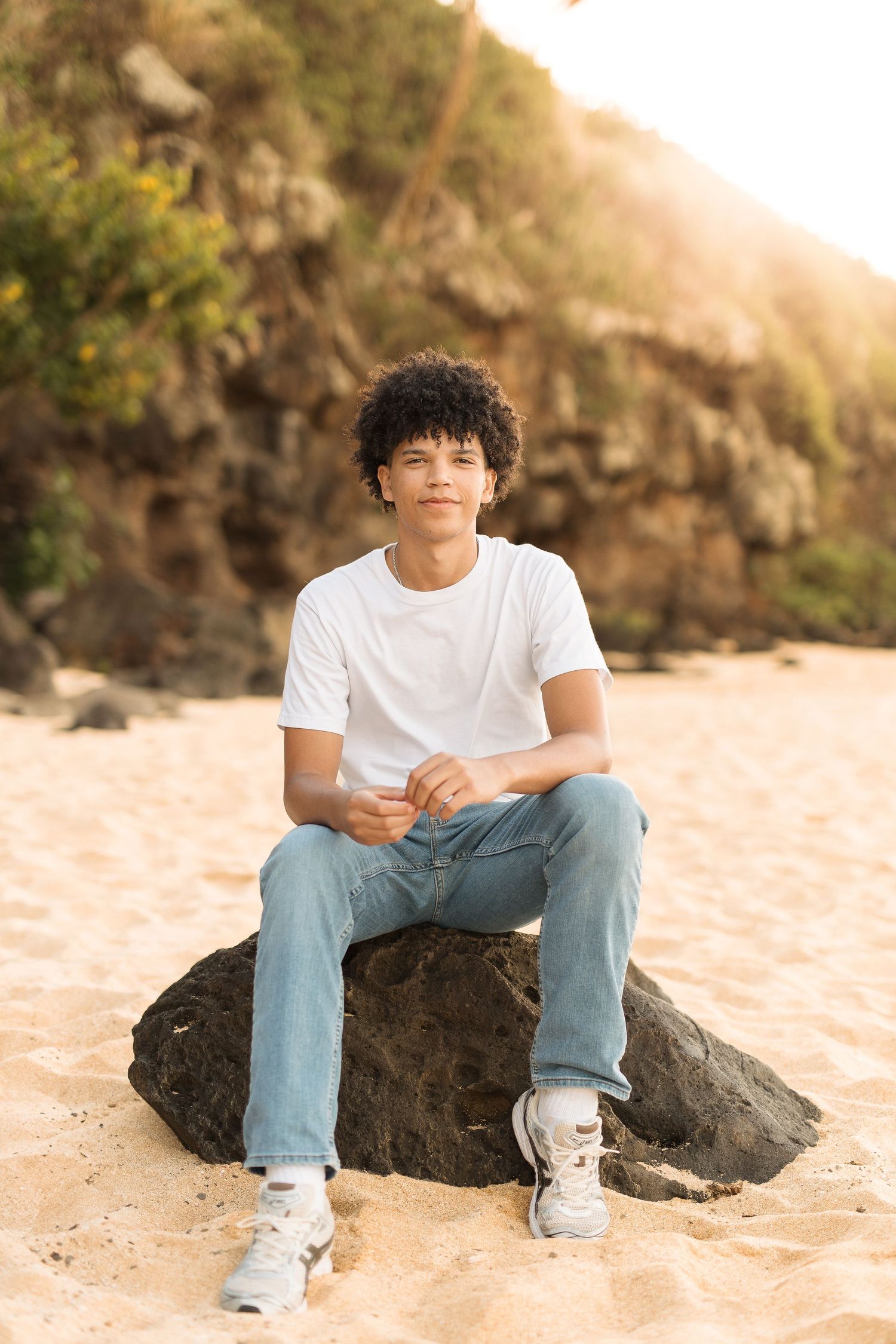 A young person in casual attire sits on a rock at the beach during golden hour.