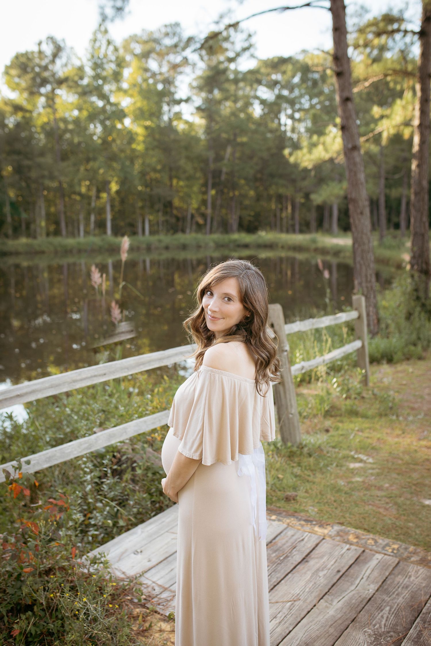 A maternity portrait with a woman in a flowing dress posing on a wooden deck by a peaceful lake.