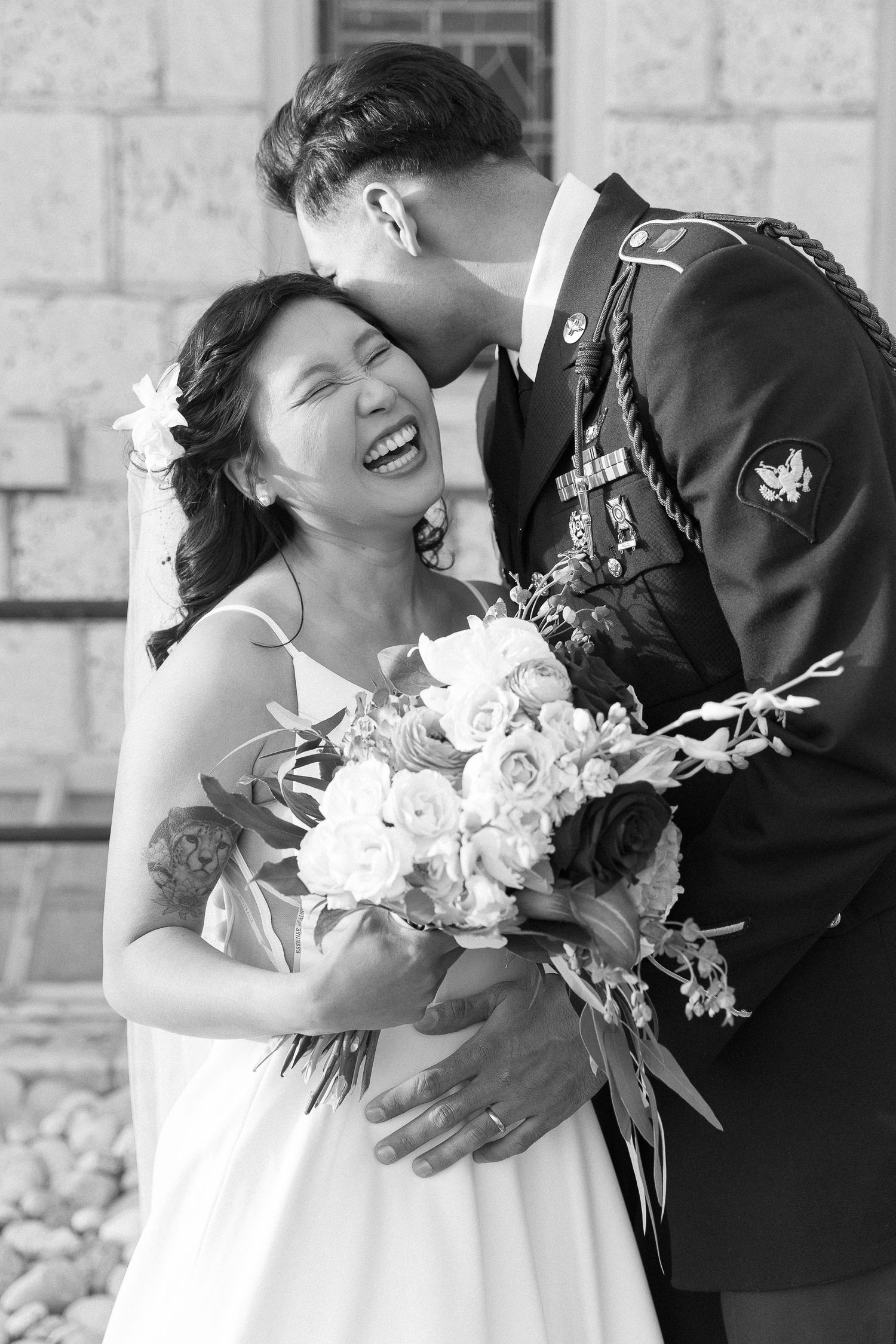 Newlywed couple share joyful laughter during outdoor wedding photoshoot with white bouquet.