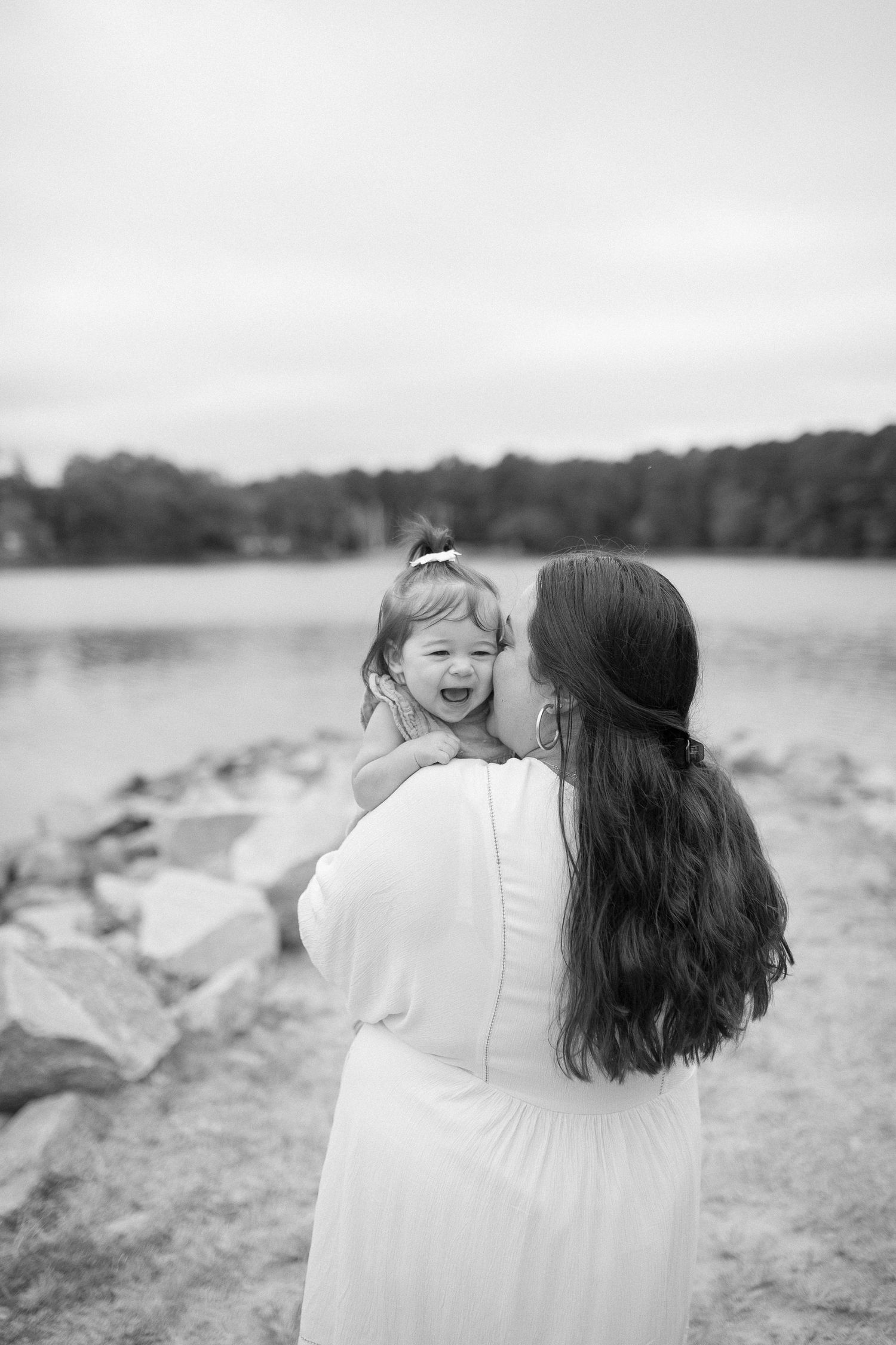Mother and baby share a joyful moment on a rocky lakeshore pathway in black and white.