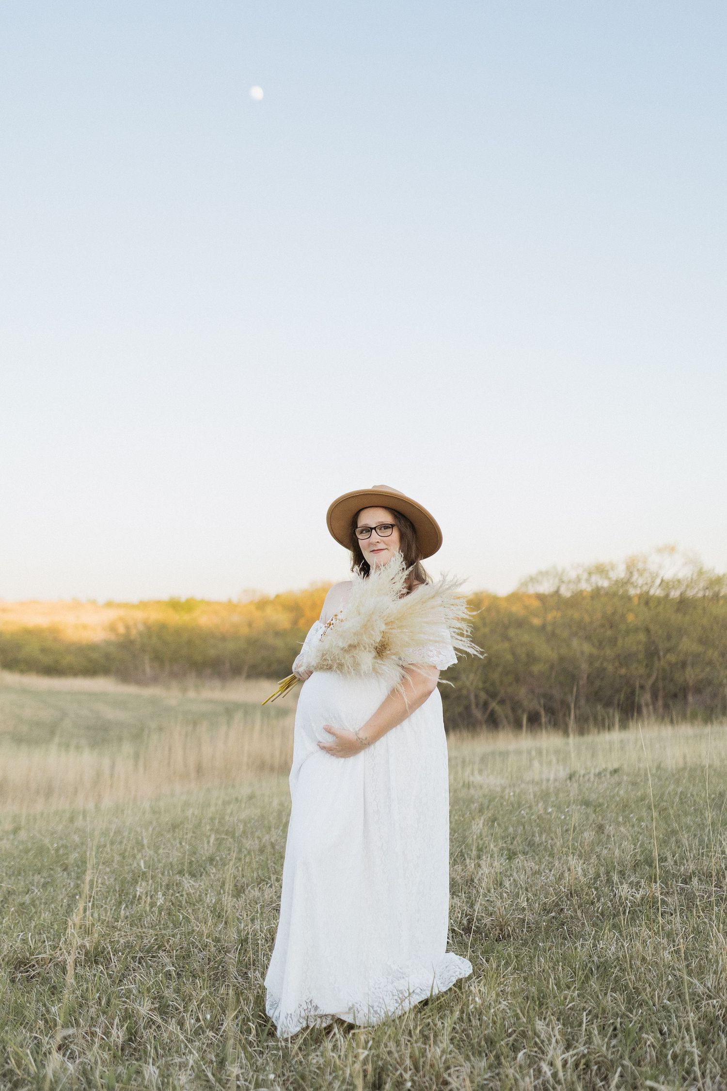 Maternity portrait in a white lace dress and hat standing in a golden field at sunset.