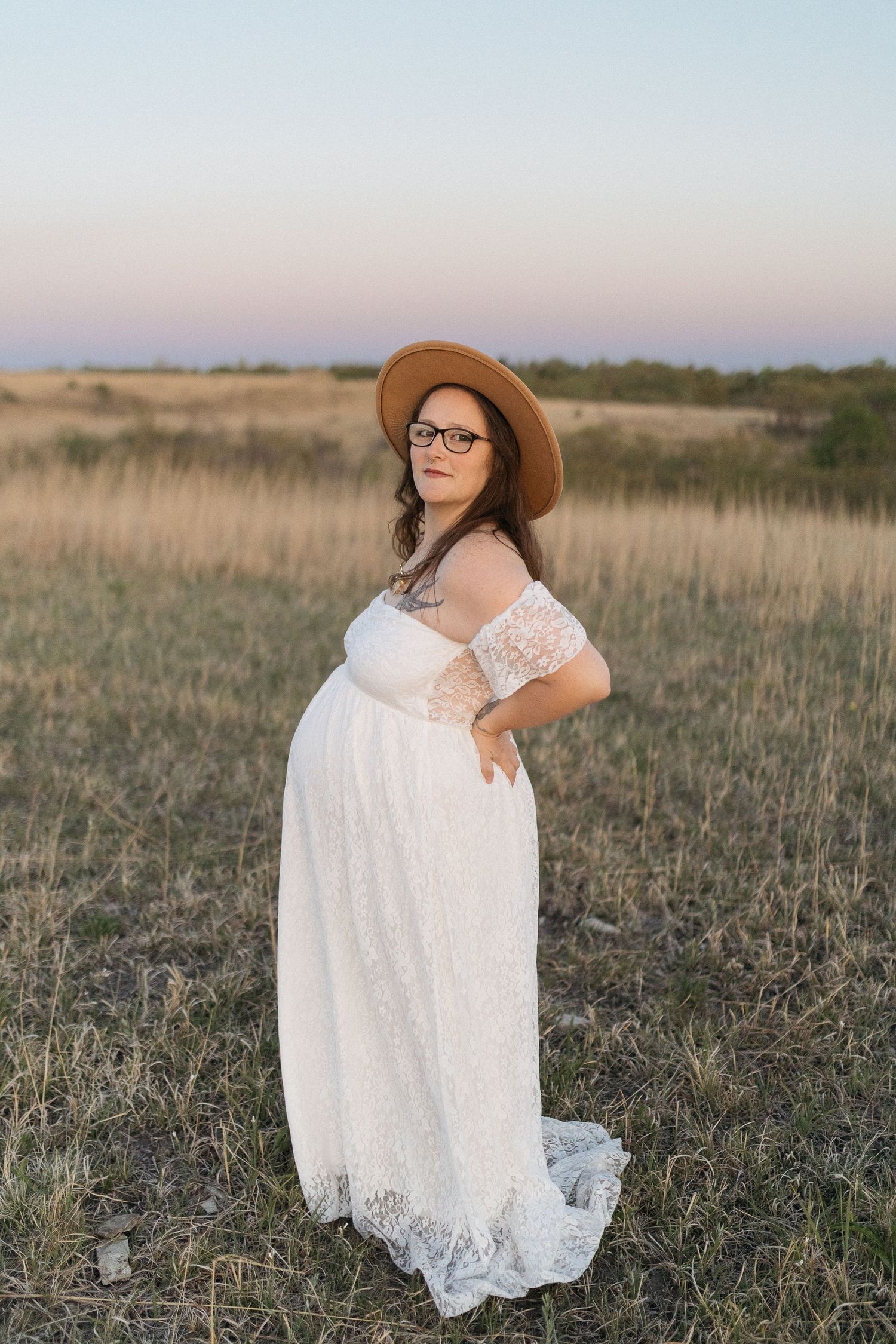 A maternity photo of someone in a flowing white lace dress and brown hat standing in a field at dusk.